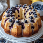 A golden-brown Lemon Blueberry Pound Cake topped with a shiny lemon glaze, garnished with fresh blueberries and lemon slices, on a white plate.